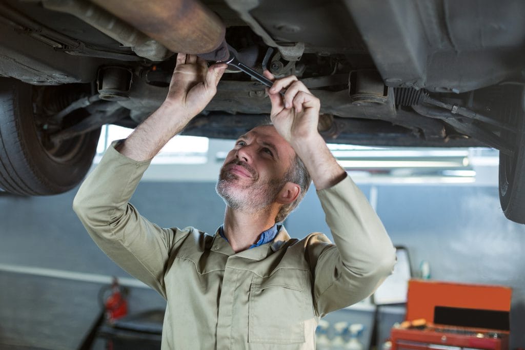 A mechanic in coveralls works under a raised vehicle, using a wrench to repair or inspect the car's underside in an auto repair shop.