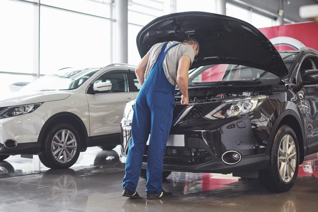 A mechanic in blue overalls inspects the engine of a black car with its hood open inside an auto repair showroom near 30A Beach. A white car is parked nearby.