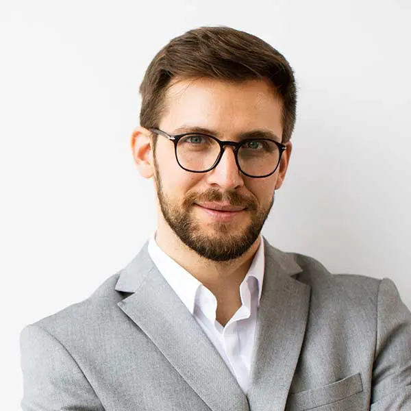 A man with short brown hair, a beard, and glasses, wearing a light gray suit and white shirt, stands in front of a plain white background—ready to represent his Panama City Beach auto repair business.