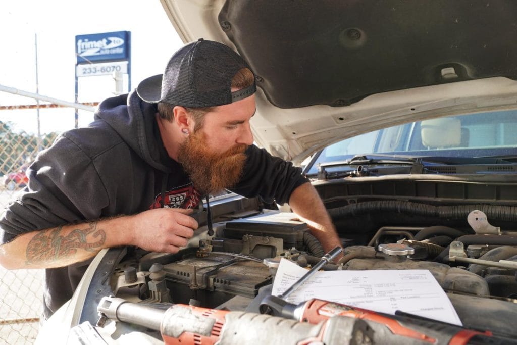 A mechanic with a beard inspects a car engine outdoors at a car repair shop in Panama City Beach, with tools and paperwork on the engine bay and a business sign visible in the background.