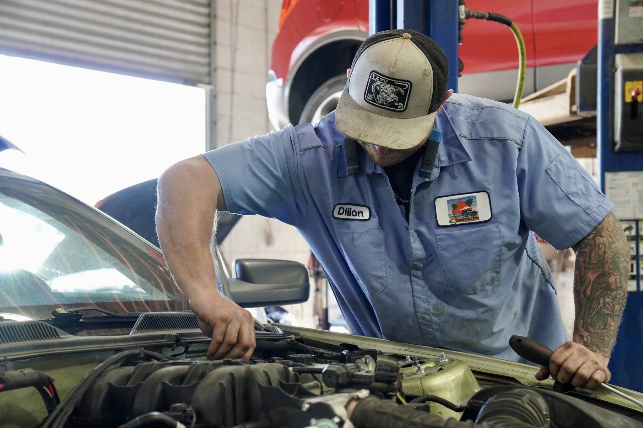 A mechanic in a blue uniform and cap works under the hood of a car in a busy car repair shop.