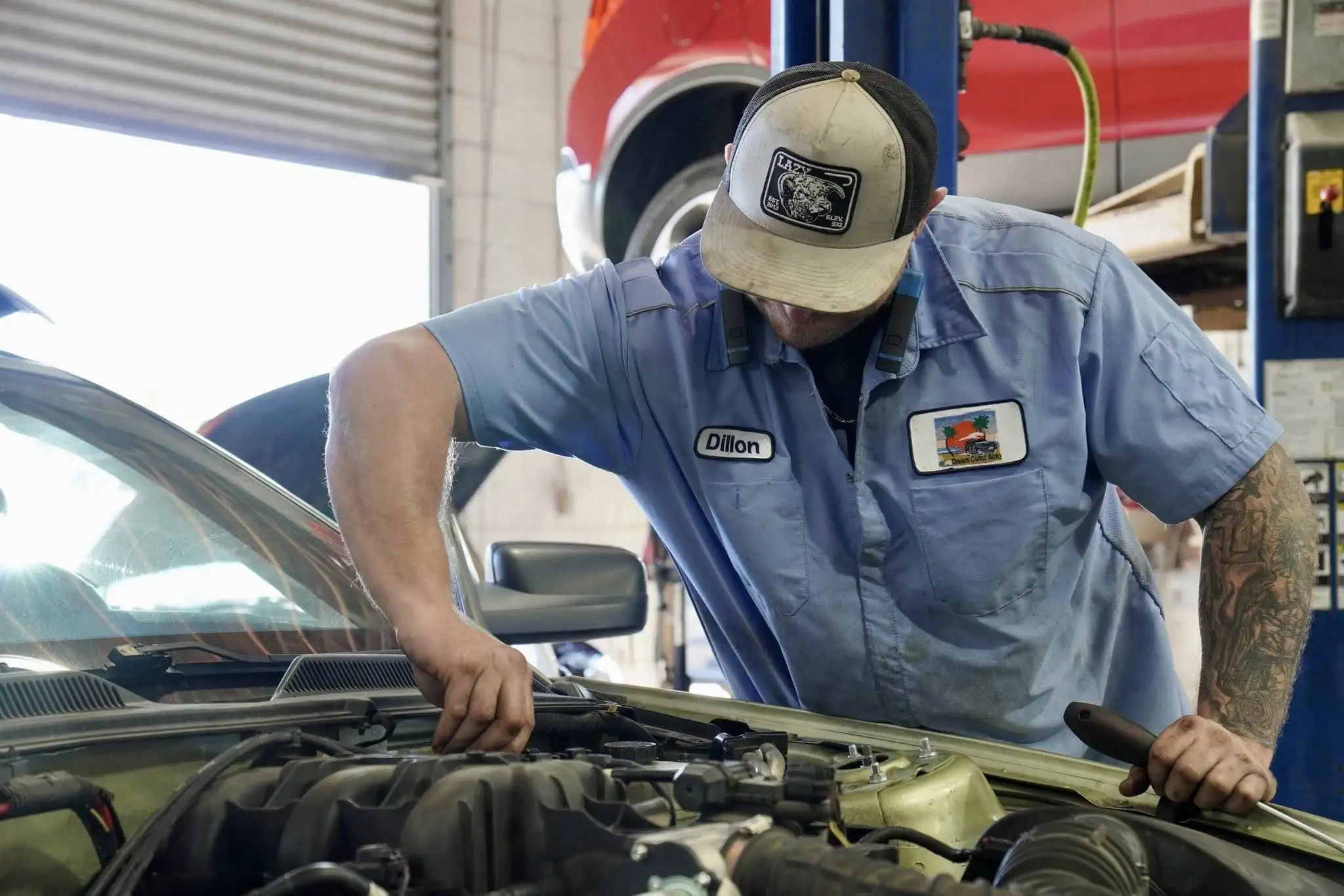 A mechanic in a blue uniform and cap works under the hood of a car in a busy car repair shop.