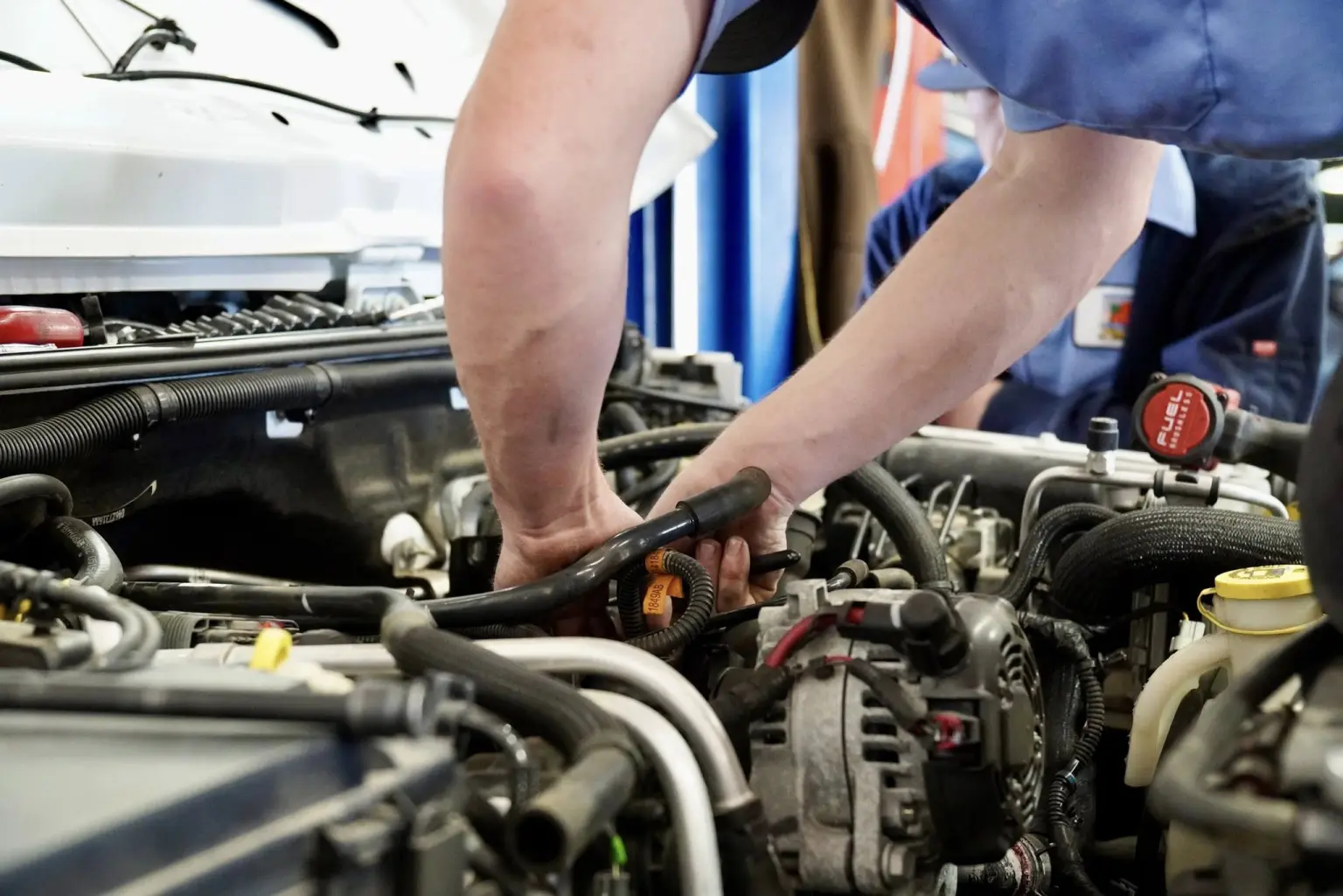 A person is working under the hood of a car, inspecting or repairing engine components with both hands at an auto repair shop.