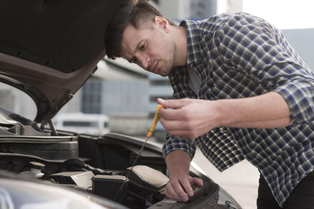A mechanic checks the oil level of a car engine using a dipstick, with the car hood open in an outdoor setting near 30A
