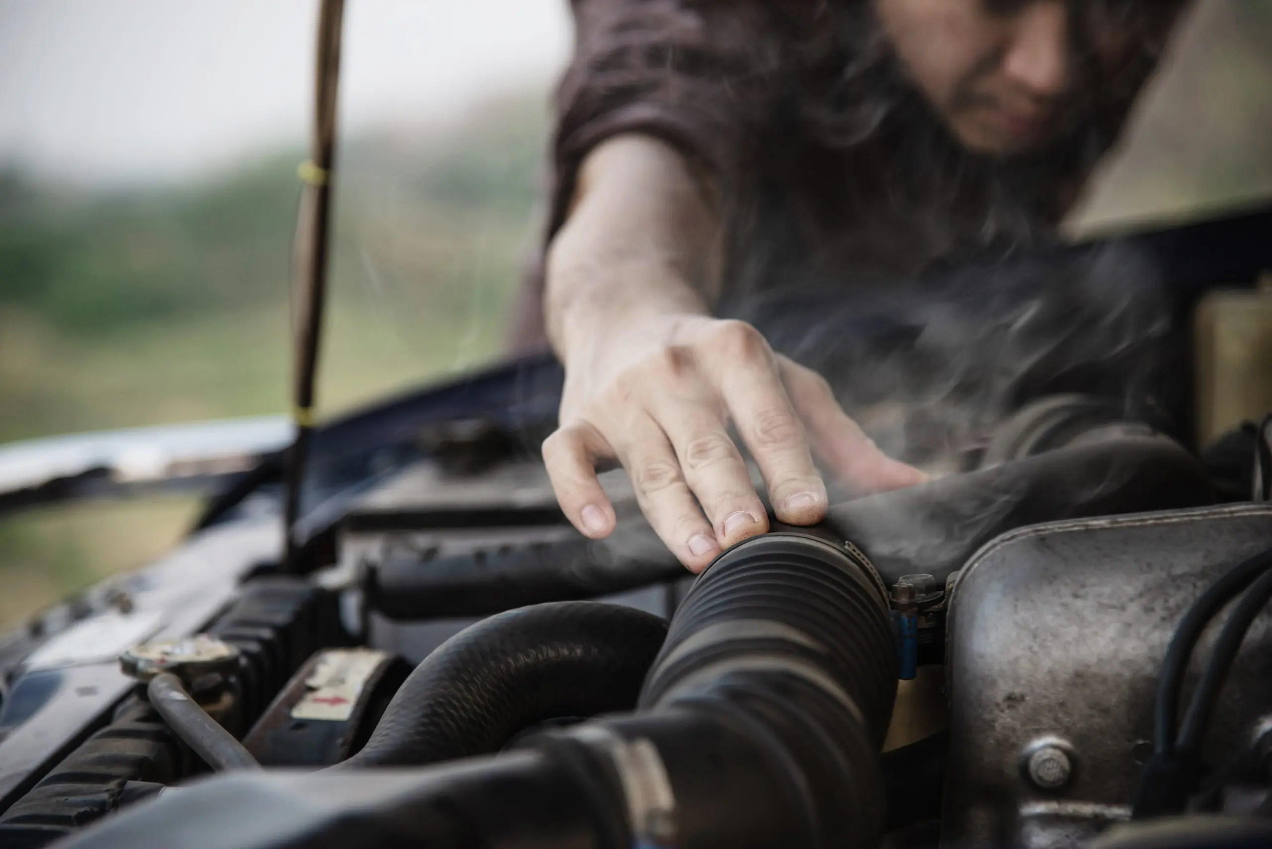 A person examines a steaming car engine under the hood, reaching out with one hand toward the vehicle's engine components—perhaps considering a visit to an auto repair shop in Panama City Beach.