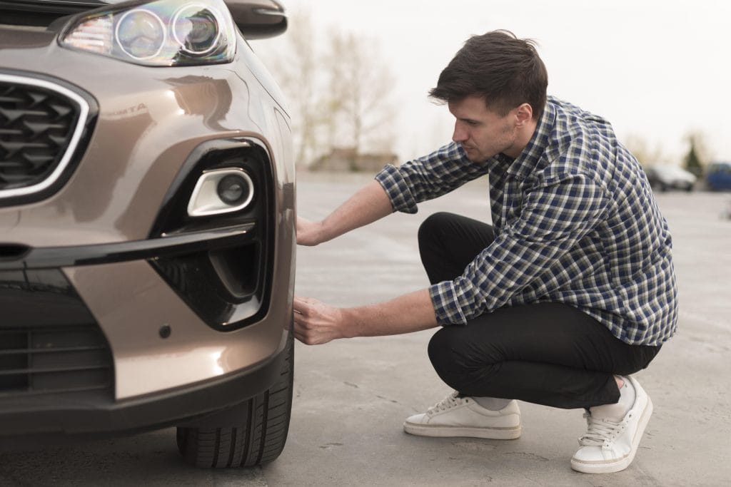 A man crouches next to a parked car in Panama City Beach, checking the front tire on an outdoor concrete surface, as though preparing for an auto repair.