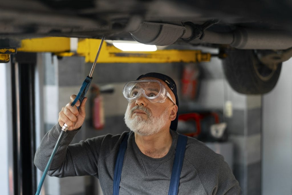 A mechanic at a Alys Beach car repair shop, wearing safety goggles, inspects the underside of a raised vehicle with a handheld tool in the garage.