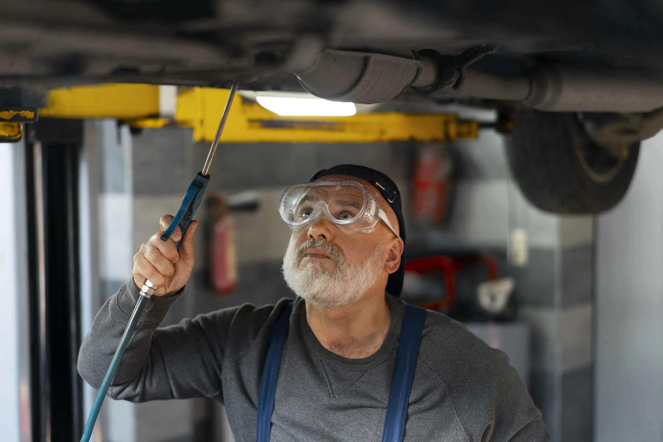 A mechanic at a Alys Beach car repair shop, wearing safety goggles, inspects the underside of a raised vehicle with a handheld tool in the garage.
