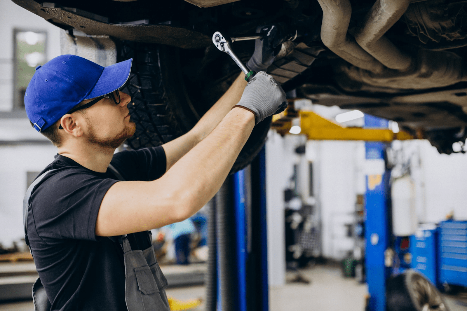 Auto technician using a ratchet under a lifted vehicle in a repair shop.