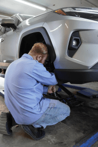 Mechanic kneeling beside a raised white SUV, working near the front wheel in a garage.  Image 2: Auto technician using a ratchet under a lifted vehicle in a repair shop.