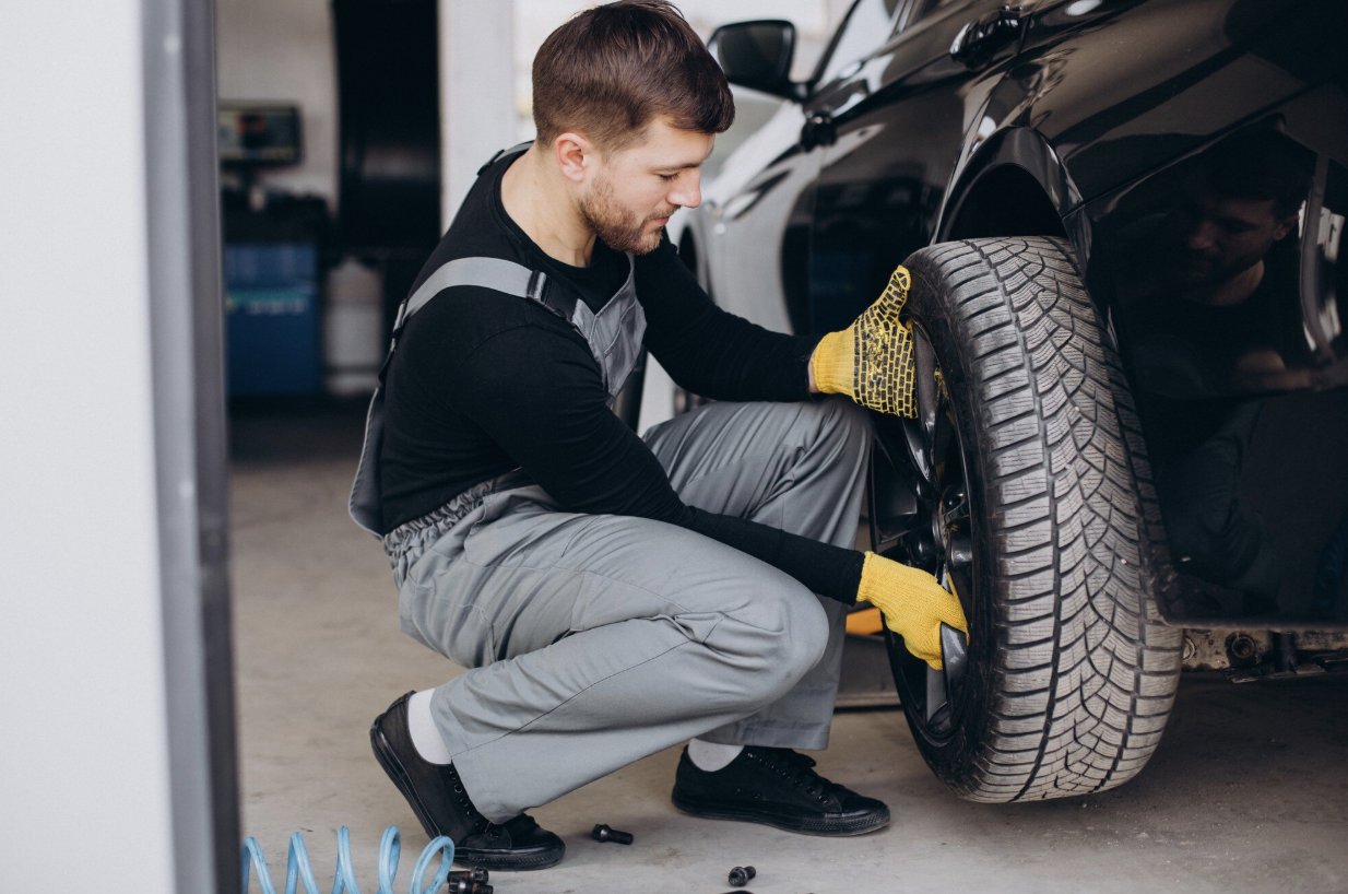 A mechanic wearing yellow gloves kneels beside a black car while changing a tire in a garage, ensuring proper tire rotation to help extend tire life.