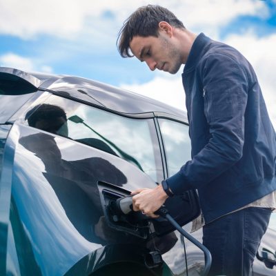 A man stands beside an electric car in Panama City Beach, plugging in a charging cable on a bright day with a blue sky and some clouds, highlighting the city's shift from traditional auto repair to greener alternatives.