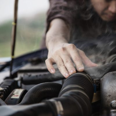 A person examines a steaming car engine under the hood, reaching out with one hand toward the vehicle's engine components—perhaps considering a visit to an auto repair shop in Panama City Beach.