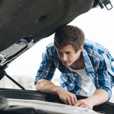 A man in a plaid shirt looks under the hood of a car at a Panama City Beach auto repair shop, inspecting the engine.