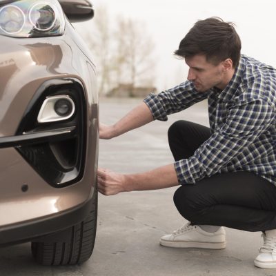A man crouches next to a parked car in Panama City Beach, checking the front tire on an outdoor concrete surface, as though preparing for an auto repair.