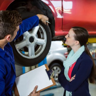 A mechanic at a car repair shop points to the rear tire area of a car on a lift while explaining an issue to a woman holding a clipboard.