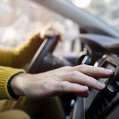 Person wearing a yellow sweater adjusts the car's dashboard controls while driving near Panama City Beach, with one hand on the steering wheel and the other on the console.