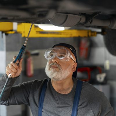 A mechanic at a Panama City Beach car repair shop, wearing safety goggles, inspects the underside of a raised vehicle with a handheld tool in the garage.